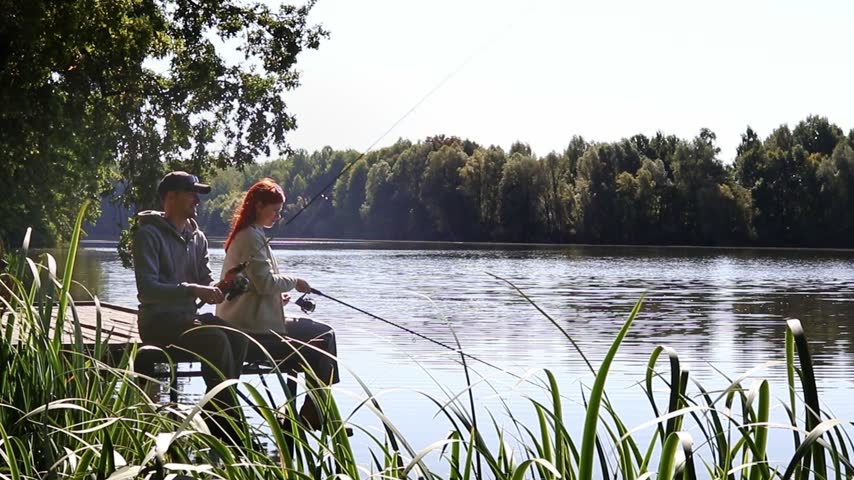 A man and a woman are sitting side by side on a wooden pier by the lake. Fishing together, relaxing together in nature and peaceful leisure by the water.
