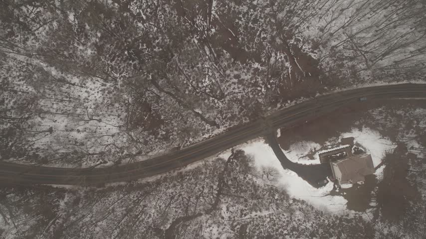 A top down perspective captures a solitary residence nestled among bare trees and patches of snow along a curved asphalt path during the cold season