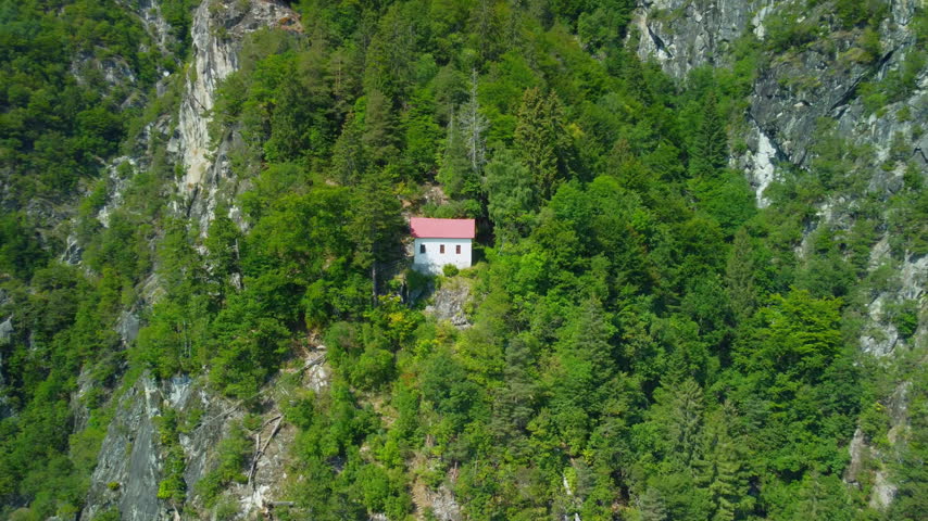 Drone mountain cabin forest cliff aerial pan right shot moving across a solitary white house with red roof nestled on a steep rocky mountainside under bright summer daylight.