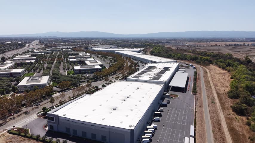 Aerial perspective of a large modern logistics warehouse with multiple delivery vans parked at loading docks next to a corporate office park complex in Milpitas, California on a sunny day