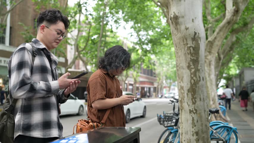 Two men stand near a street tree and look down at smartphones on a city sidewalk.