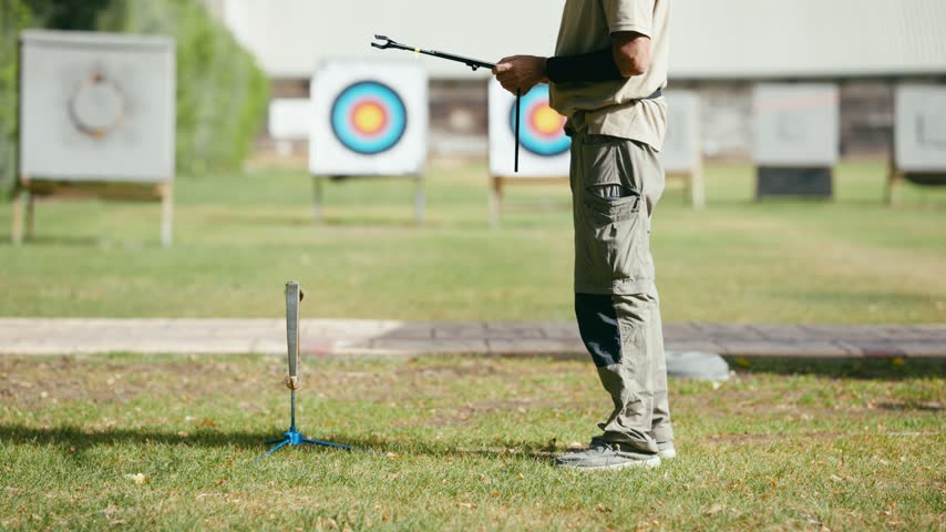 Senior man placing his bow on a stand after practicing archery. Archer finishing his training on a green field with targets in the background