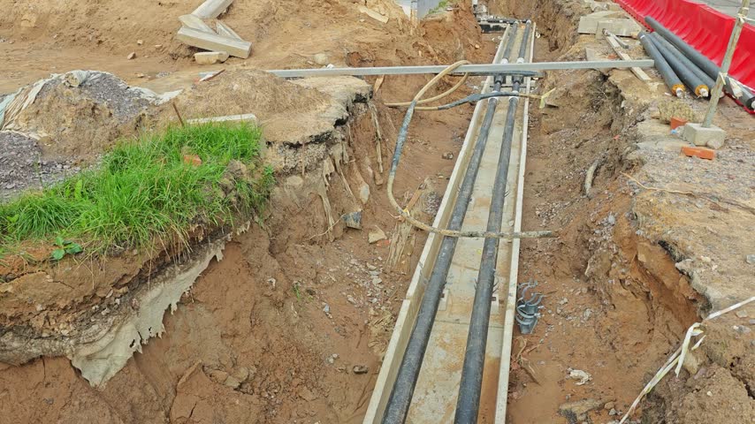 Construction trench with underground cable conduits in concrete trays; exposed earth walls, red plastic barrier, scattered construction debris, utility pipes and clamps