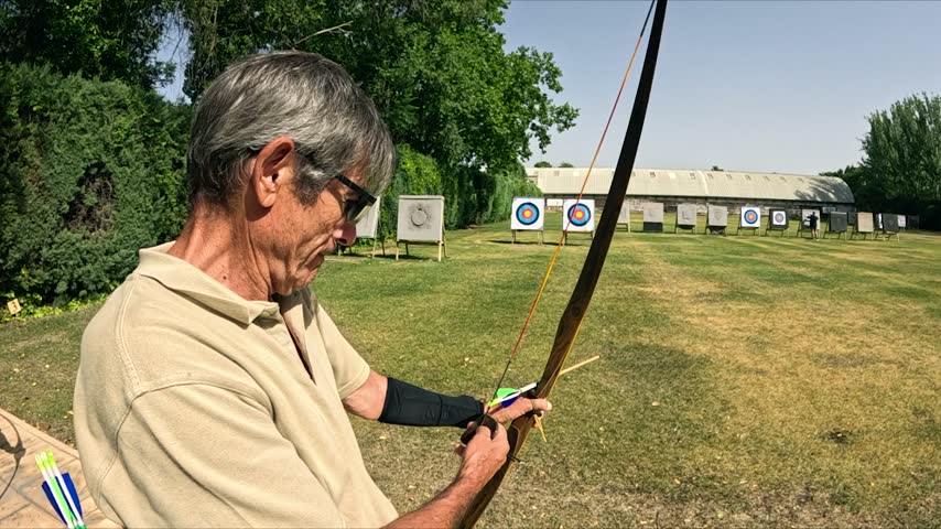 Concentrated senior archer practicing at an outdoor range. He nocks an arrow, draws the string, aims, and shoots his traditional longbow