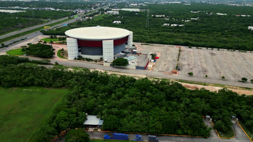 Aerial view circling a large modern convention center or arena, revealing its unique circular architecture surrounded by a vast parking lot, lush green jungle and a busy highway in the background