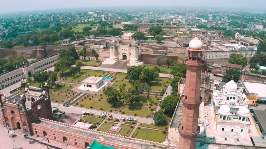 High-angle panorama showing Alamgiri Gate at Lahore Fort, a Badshahi Mosque minaret, the white Samadhi of Ranjit Singh, and the central Hazuri Bagh Baradari garden in Lahore, Pakistan.