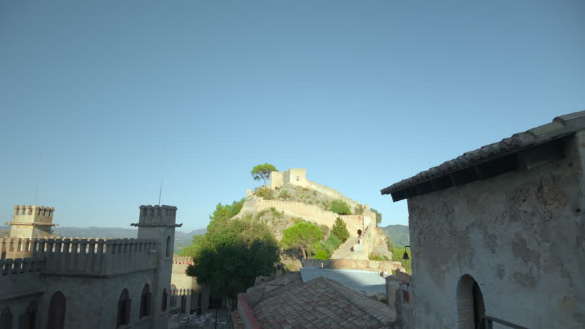 Historic hilltop fortress framed by rough stone buildings with tiled roofs and clear blue sky, warm sunlight on central walls, deep foreground shadow, layered architecture