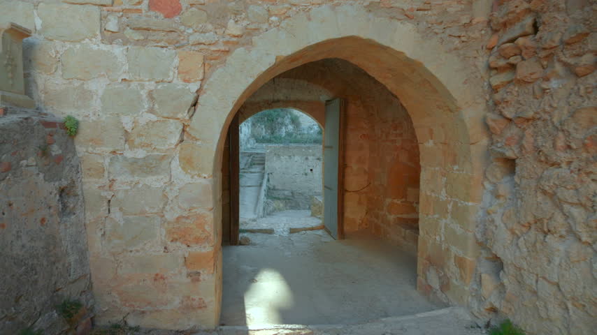 Weathered stone passage opening from dark interior into bright fortress walkway with centered arch, rough cobblestone floor, and aged cliffside masonry under soft daylight