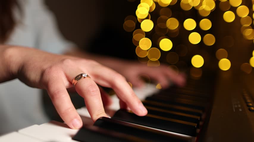Slow motion camera pulls back from female hands playing piano in festive setting. Golden bokeh reflections spread across keys creating spacious warm atmosphere.