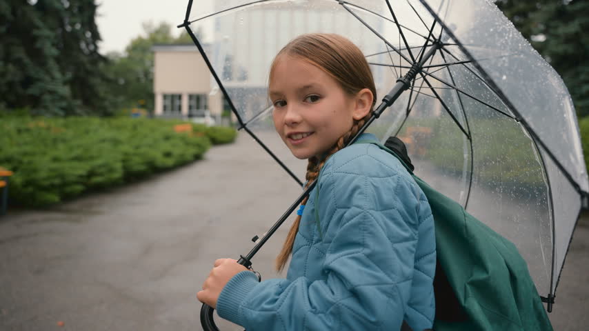 Back view Caucasian little school girl walking at rainy city holding umbrella child turning around smiling looking at camera kid daughter pupil schoolgirl with parasol rotate smile walk to studying