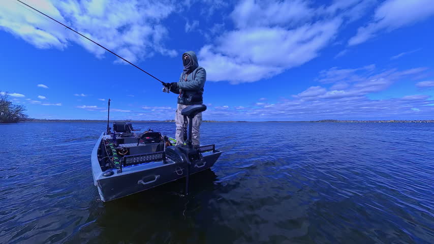Fisherman fishing on lake from boat casting spinning rod at daylight. Angler is standing in an equipped boat and catching fish while trolling. Man holds spinning rod while offshore during sunny day