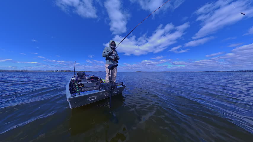 Fisherman fishing on lake from boat casting spinning rod at daylight. Angler is standing in an equipped boat and catching fish while trolling. Man holds spinning rod while offshore during sunny day