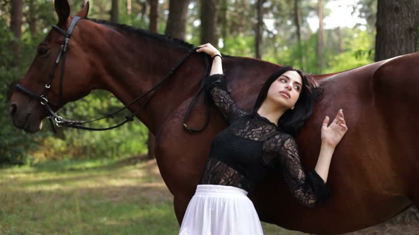 A woman in a white dress posing with a brown horse in the forest at sunset, a symbol of harmony with nature. Sexy girl holds dark brown stallion by reins.