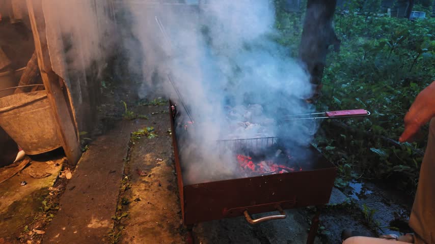 Delicious pieces of meat cooking on a hot charcoal brazier grill during a backyard barbecue party at dusk, with smoke billowing from the glowing embers as a man tends to the food