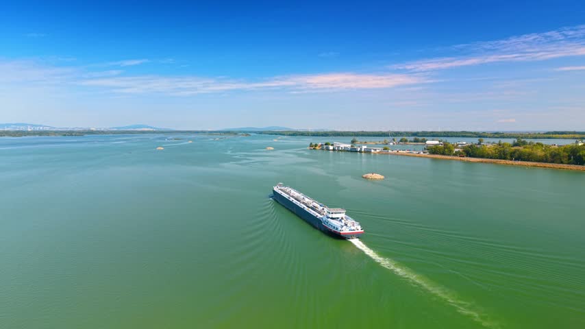 Barge cruising on green reservoir waterway. Aerial angle shows freight boat sailing forward on blue water body with distant shore.