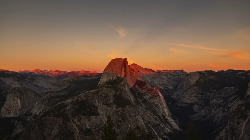 A wide panoramic view from Glacier Point capturing the iconic granite monolith of Half Dome surrounded by the High Sierra mountain peaks during a vibrant golden hour sunset in Yosemite National Park, California.