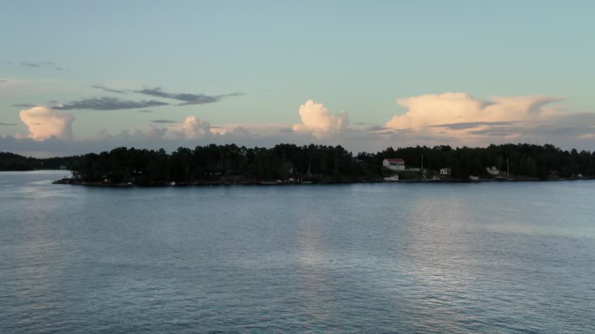 Wide view of a Swedish archipelago island with white waterfront houses, dense forest and calm water beneath towering clouds at dusk.