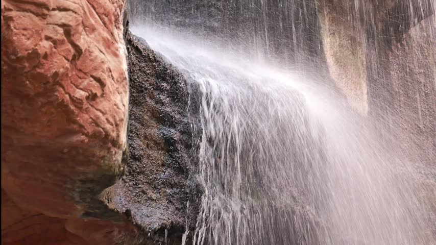 A red rock waterfall cascades over a sandstone cliff, creating misty spray and silky flowing water against textured rock layers in a quiet canyon setting, evoking nature, motion, and erosion