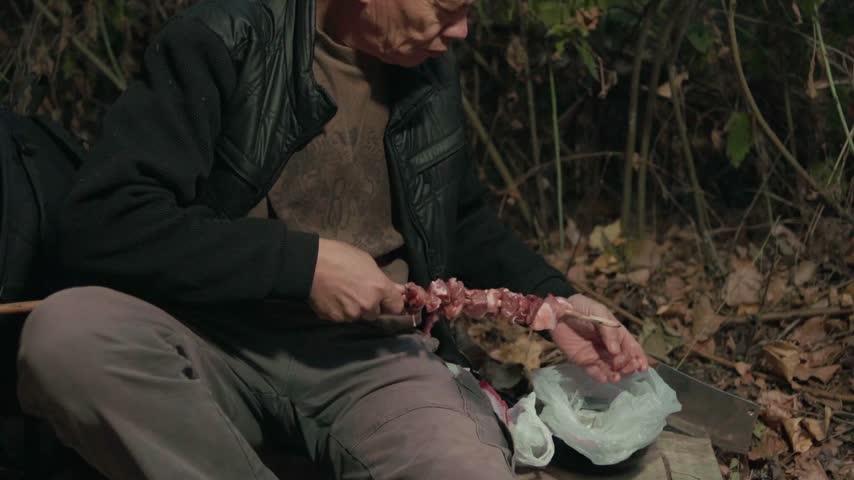 Mature man threading raw meat pieces onto wooden skewer in outdoor camp.