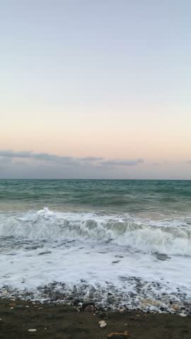 Sea waves crash with white foam over rocky pebble beach during calm evening twilight. Slow motion vertical footage