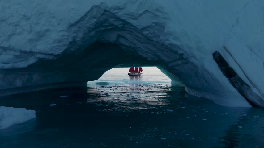 Aerial view of a red-sailed ship framed by a natural ice arch in the arctic waters of Greenland.