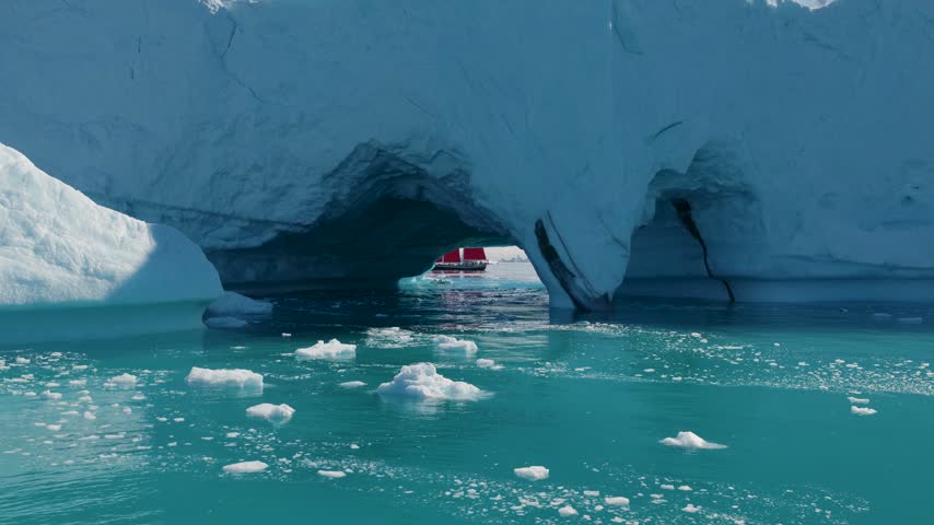 Aerial view of a red boat sailing through a natural arch in a massive blue iceberg floating in the cold waters of Greenland.