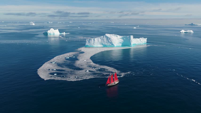 Aerial view of a red-sailed ship navigating through calm blue waters near a large white iceberg in Greenland.