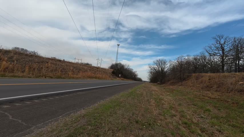 Stationary roadside footage filmed along Historic Route 66 in Oklahoma during early spring. Vehicles pass along a rural two-lane highway surrounded by grassy embankments, leafless trees and utility poles beneath a partly cloudy sky.