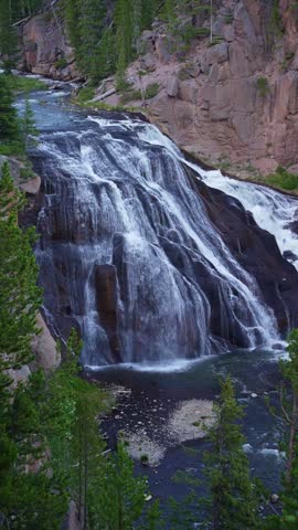 half speed vertical clip of Gibbon Falls in Yellowstone National Park cascades down rocky cliff face. Mist rises from pool, swirling with spray. Rugged cliffside frames the falling water