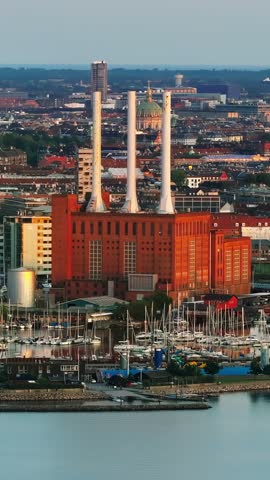 Aerial view showing historic power plant brick building with three tall chimneys near harbor featuring docked boats and city skyline background.