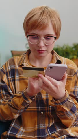 Woman using credit bank card and smartphone while transferring money, purchases online shopping cashless, order food delivery at home apartment indoors. Happy young lady sitting in room on couch