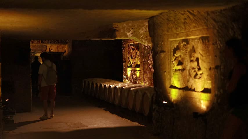 Visitors walking through dimly lit historical stone cellar featuring ornate bas relief carvings on rock walls and rows of stacked wine barrels in subterranean cave atmosphere.