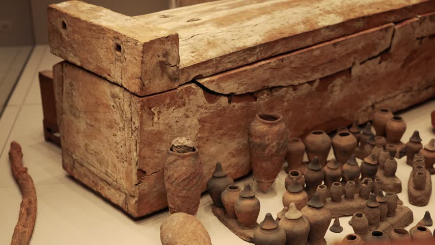 Historical display featuring antique wooden coffins alongside diverse collection of clay pots and small ceramic burial goods positioned on museum shelves under professional soft studio lighting.