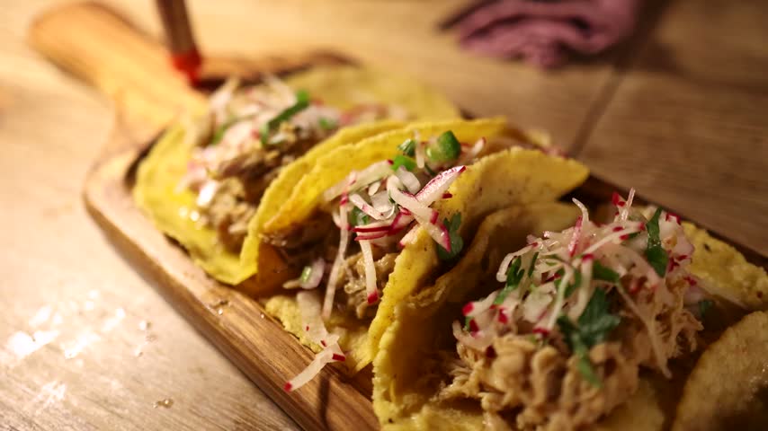 Close up of hands arranging gourmet tacos with shredded meat and fresh garnishes on a rustic wooden serving board.