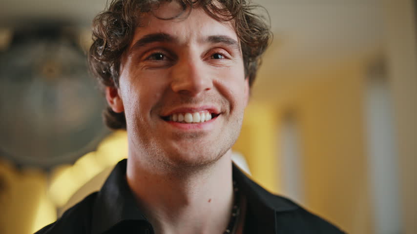 Young man raising DSLR camera aiming lens directly at viewer, close up portrait. Curly hair photographer shooting indoors holding professional camera, creative photography passion enthusiast hobby.
