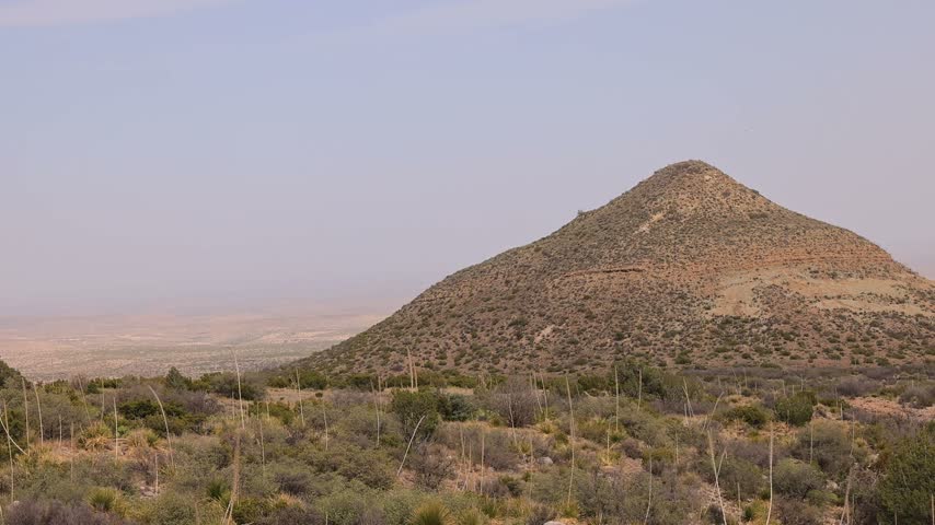A static wide shot captures a large, cone-shaped hill covered in sparse vegetation rising from an arid desert environment under a hazy sky.