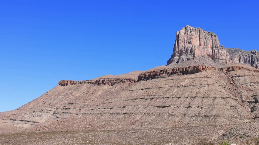 A massive red rock butte rises above a sloping desert hill with visible sedimentary layers under a clear blue sky.