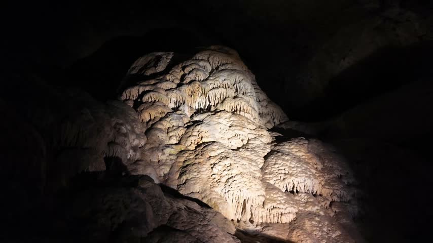 A large, textured stalagmite rises from the cave floor, illuminated by a focused light beam against a pitch-black background.