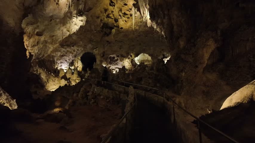 A group of people walk along a lit pathway with railings through a large cavern featuring stalactites and rock formations.