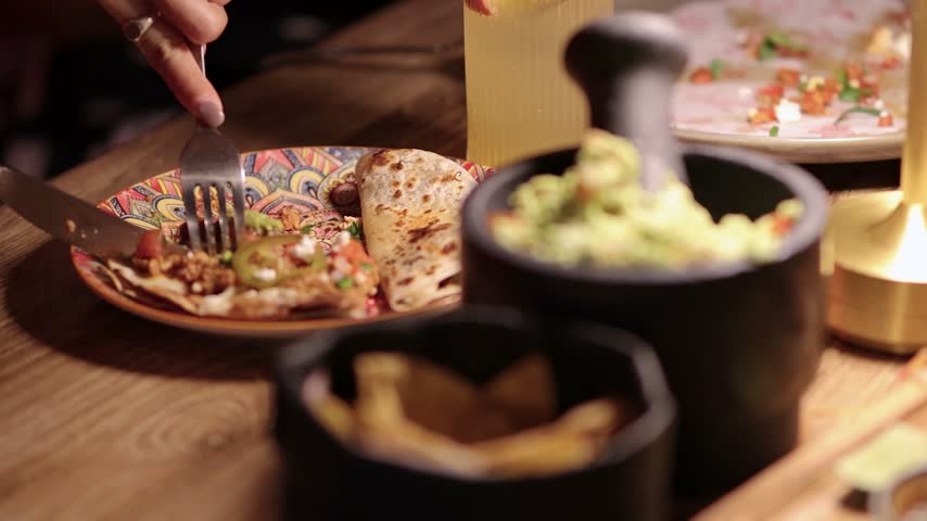 Group of friends dining at mexican restaurant featuring close up shot of dipping tortilla chip into bowl of fresh guacamole served in stone mortar while quesadillas are prepared nearby.