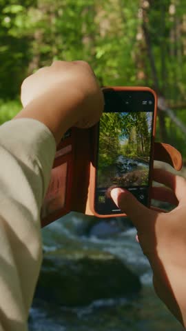 Hand holds phone, capturing forest stream in warm sunlight. Person focuses, adjusting frame for perfect shot. Nature photography, outdoor adventure, travel vlogging, serene woodland moments