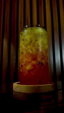 Dramatic low angle zoom-in shot of iced pineapple fruit tea layered with red syrup, showcasing a frosty glass with water droplets on a teak wood table under warm, dim studio light.