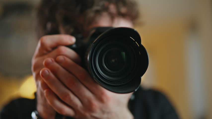 Photographer holding DSLR camera pointing lens toward viewer, then lowering it revealing cheerful smiling face. Close up hands gripping professional camera gear, creative hobby home photo studio.