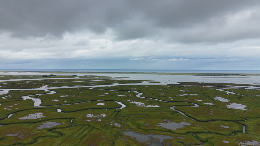 Smooth drone fly-over of curving water channels pattern Ipswich coastal marsh beneath a heavy overcast sky with the open sea visible along the distant horizon, Massachusetts, United States