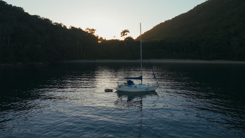 Aerial view of the sailing boat anchored in the calm lagoon with sun rising and shining from the mountains