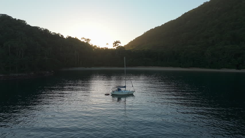 Aerial view of the sailing boat anchored in the calm lagoon with sun rising and shining from the mountains