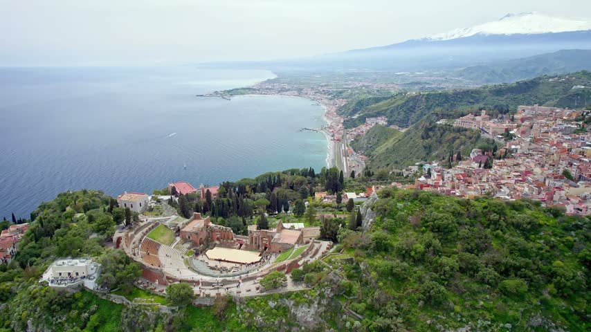 A Gentle Aerial Arc Holds Teatro Antico di Taormina in the Foreground While the Coastal Town, Ionian Sea and Snowcapped Mount Etna Fill the Distance.