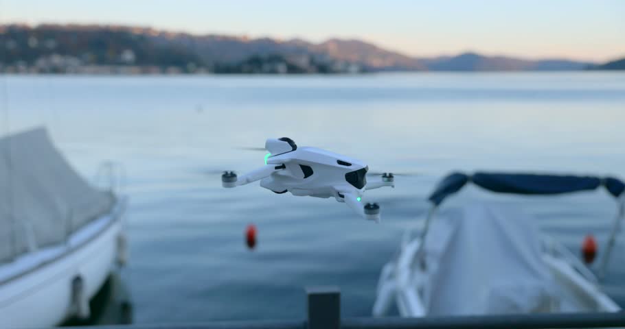 Close up of a compact white drone hovering in mid-air over a lake with boats at sunset