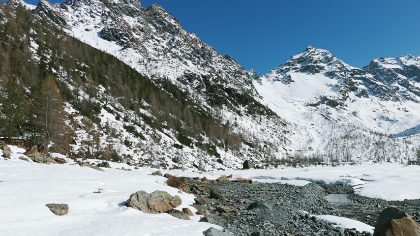 Man in a red jacket walking along a snowy path next to a rocky mountain stream in Alpe Ventina, Italy. Aerial backward