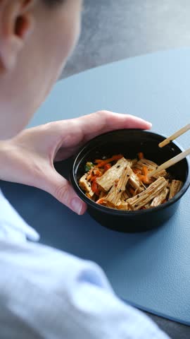 Close up person eating healthy asian takeaway salad with yuba, carrots, and sesame seeds from black plastic bowl using wooden chopsticks during quick office lunch break. Woman eating asian yuba salad